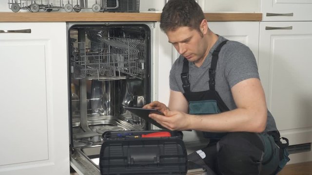 Handyman with tablet pc repairing domestic dishwasher in the kitchen.