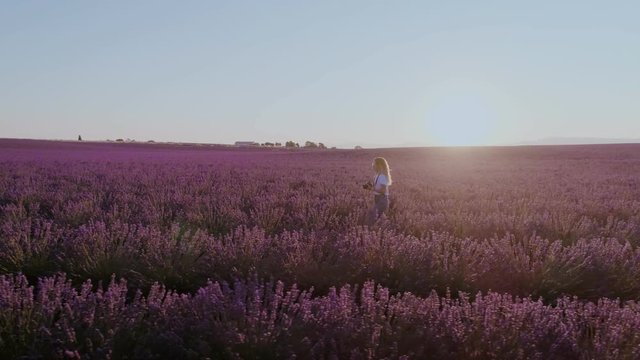 Young beautiful woman in millennial hipster outfit, freelance photographer or social media internet influencer or blogger, walks with camera through dreamy puple lavender fields. content creator nomad