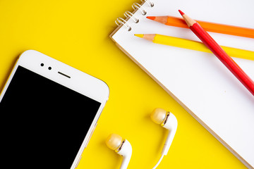 White smart phone and headphones with notepad and pencils on the yellow background.