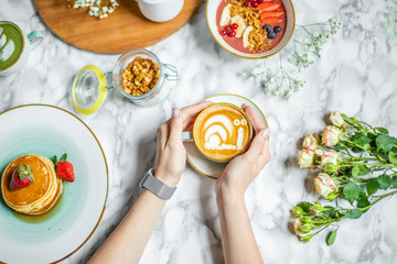 a cup of coffee with milk in hand, pancakes and cereal with fruit on the table
