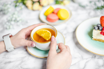 a cup of tea in the hands of a girl, on the table cheesecake, macaroon and flowers