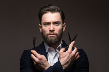 Portrait of bearded hipster male barber holds sharp scissors and a blade isolated on gray backgrund