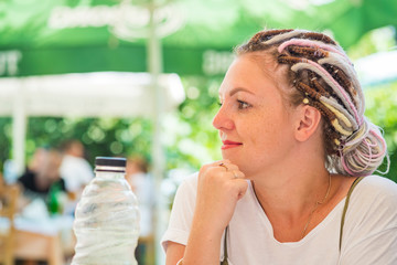 Cute young girl with freckles and colorful dreadlocks is sitting at a table in an open-air cafe, next to her is a plastic bottle of water. Close-up portrait of a girl smiling
