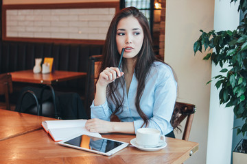 Beautiful charming brunette smiling Asian girl working or studying at table in cafe, student,...