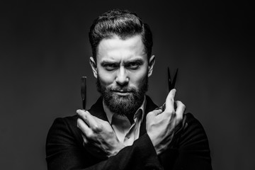 Portrait of bearded hipster male barber holds sharp scissors and a blade isolated on gray backgrund