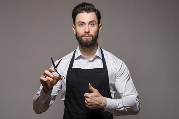 Portrait of bearded barber holding equipments in hand, looking at camera, isolated on gray background