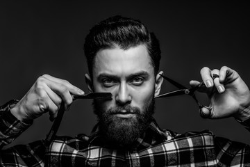 Handsome bearded young man in a plaid shirt holding a sharp professional scissors and razor isolated on dark background. Close-up