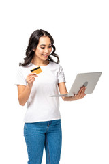 Smiling pretty asian woman in white t-shirt and blue jeans using laptop and holding credit card isolated on white