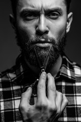 Fototapeta na wymiar Portrait of stylish professional hairdresser with beard wearing shirt holding scissors near his beard on gray background