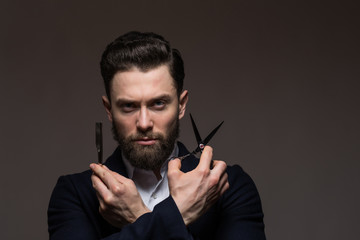 Bearded young man caucasian hipster with moustache holding scissors and straight razor on gray background. Mens haircut, shaving. Male in barbershop.