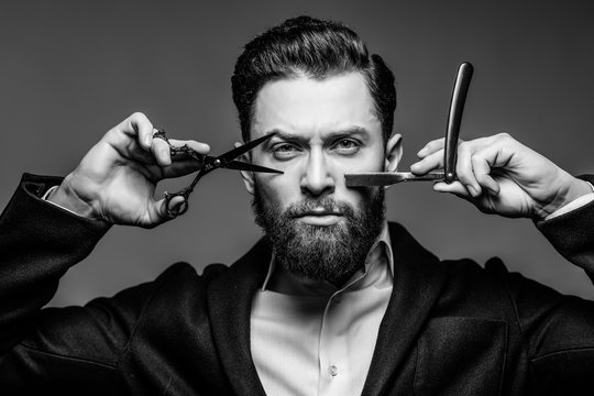 Portrait of young bearded man in suit holding barber scissors and straight razor standing isolated on gray background