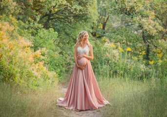 Obraz na płótnie Canvas young sweet pregnant girl with light curly hair hugs and holds her tummy, standing in a fairy forest in cold shades. dresses a long elegant satin silk pink dress with a lace top. art processing photo