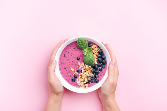 Woman's hands holding blueberries smoothie bowl with mint on pink background. Top view, copy space.