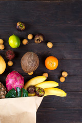 Paper bag of different healthy tropical fruits on dark wooden background. Top view.