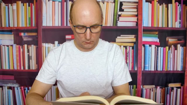 Serious man in glasses reading book at table in home office on bookshelf background. Man professor preparing for classes in college library on bookcase background thinking thoughtful man