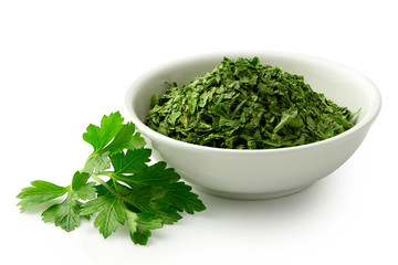 Dried chopped parsley in white ceramic bowl next to fresh parsley leaves isolated on white.