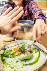 A hungry girl in a plaid shirt with a knife and fork tries to cut off a piece of food in the plate of his young man, but he does not allow this to be done with a gesture of his hand. Close-up.