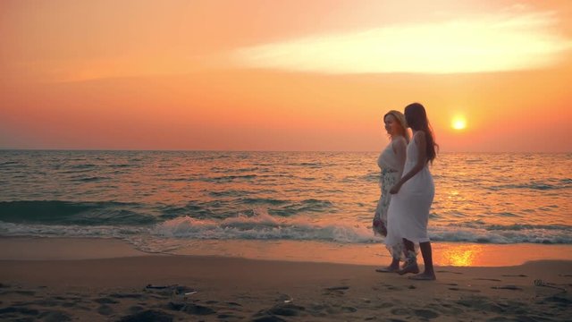 mother and daughter in white dresses walk barefoot on a sandy beach, holding hands against the backdrop of a magnificent sunset