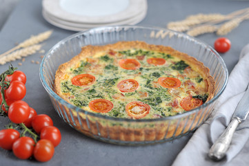 Classic French quiche pie with bacon and spinach. In the frame of cherry tomatoes, plates, a knife, a napkin made of cloth, ears of wheat. Gray background. Close-up. Macro shooting.
