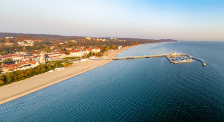 Wide aerial panorama of Sopot resort near Gdansk in Poland in sunrise light. Wooden pier (molo) with marina, yachts, beach, old lighthouse, vacation infrastructure, hotels, park and promenade - obrazy, fototapety, plakaty