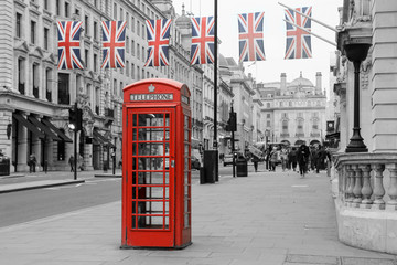 Fototapeta na wymiar A traditional British red telephone box is standing on the street. Six British flags are hanging above it. Booth and flags isolated in a black and white picture. 