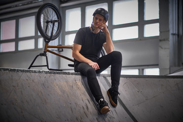Fototapeta na wymiar Pensive young man chilling after practicing tricks, sitting on a floor in a skatepark indoors