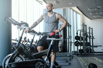 Fototapeta na wymiar Athletic man with a tattoo on his hand standing next to a exercise bike in the modern fitness club
