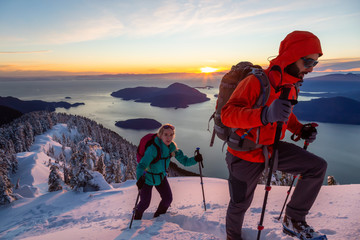 Adventure seeking man and woman are hiking to the top of a mountain during a vibrant winter sunset....