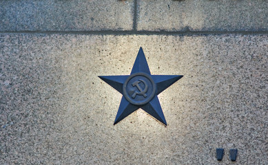 Hammer and sickle bronze star application on sunlit red granite wall.