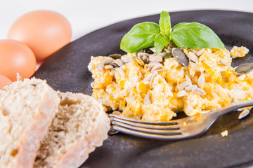 Scrambled eggs with sunflower and pumpkin seeds, some fresh eggs and wholemeal bread, eaten with a fork, decorated with basil, on a white background