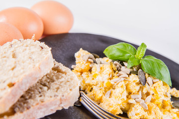 Scrambled eggs with sunflower and pumpkin seeds, some fresh eggs and wholemeal bread, eaten with a fork, decorated with basil, on a white background