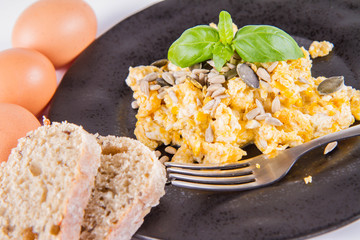 Scrambled eggs with sunflower and pumpkin seeds, some fresh eggs and wholemeal bread, eaten with a fork, decorated with basil, on a white background