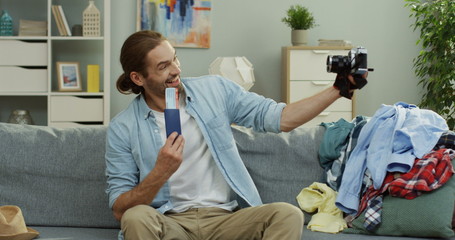 Obraz premium Attractive and cheerful young Caucasian man taking selfie photos on the camera of him holding a passport with a ticket while sitting on the sofa among mess before traveling.