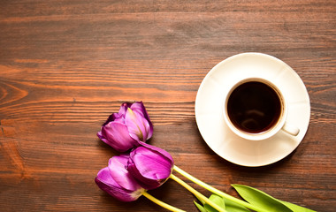 a cup of coffee or tea on a saucer stands on a wooden background, purple tulips lie next