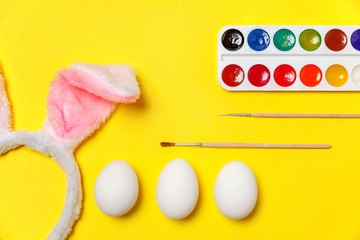 Happy Easter concept. Preparation for holiday. Decorative eggs colorful paints and bunny ears furry costume toy isolated on trendy yellow background. Simple minimalism flat lay top view copy space