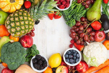 Healthy rainbow fruits vegetables berries frame, strawberries oranges plums grapes broccoli cauiliflower mango persimmon pineapple on white wooden table, top view, copy space, selective focus