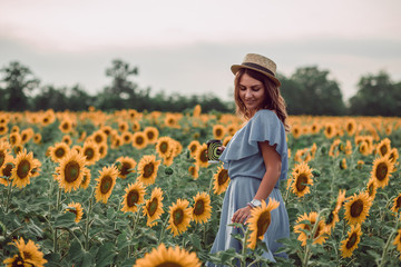 Fototapeta premium Dreaming young woman in blue dress and hat holding a cup of coffee in a field of sunflowers at summer, view from her side. Looking down