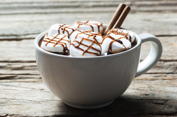 Hot chocolate with cinnamon stick and marshmallow candies with chocolate sauce in white ceramic mug or cup on wooden background. Hot cocoa and chocolate drink concept.
