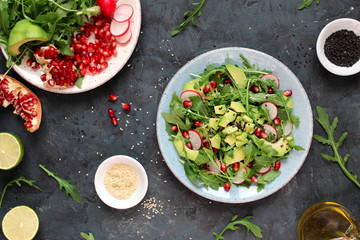 Ingrdient for making salad on dark background. Vegetable salad in plate, avocado, arugula, pomegranate, radish and sesame. Top view with copy space. Vegetarian and vegan food.