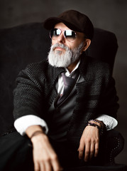 brutal senior millionaire man in classic suit white shirt and tweed jacket sitting in armchair and looks away stylish fashionable men