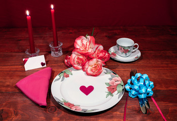 Floral pattern fine china dinnerware with matching plate, cup and saucer. bouquet of orange and white rpses, pink napkin, silverware, red candles and card