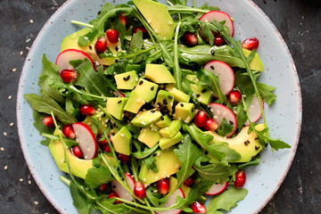 Ingredient for making salad on dark background. Vegetable salad in plate, avocado, arugula, pomegranate, radish and sesame. Top view with copy space. Vegetarian and vegan food.