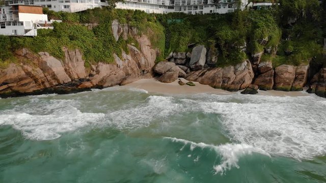 Slowly backing away from the cliff rocks of Joatinga beach in Rio de Janeiro during high tide revealing the green ocean waves rolling in