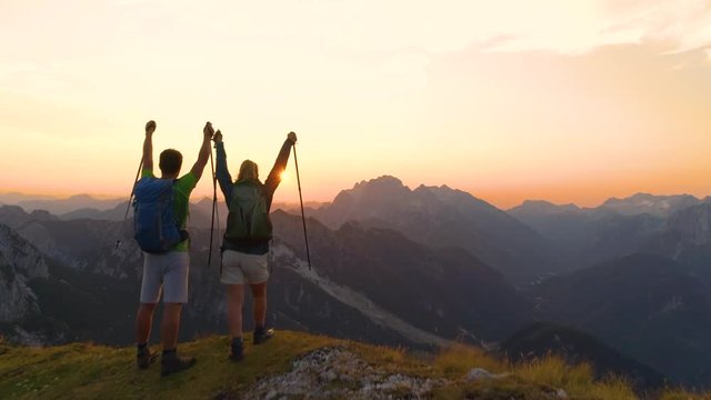 AERIAL SUN FLARE: Unrecognizable young hiker couple outstretch their arms as they catch the sunset while hiking in the Julian Alps. Picturesque shot of golden sunrise illuminating the excited couple.