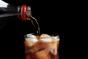 Pouring refreshing cola from bottle into glass with ice cubes on black background, closeup