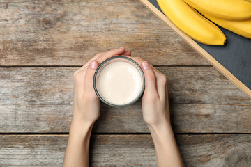 Woman holding glass of healthy protein shake near bananas at wooden table, top view