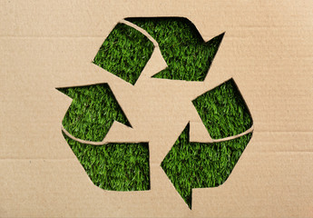 Sheet of cardboard with cutout recycling symbol on green grass, top view