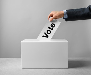 Man putting his vote into ballot box on table against light background, closeup