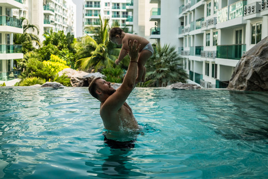 Dad Throws Baby In The Pool On The Background Of Coconut Palms. Happy Active Family Young Father And His Cute Daughter Adorable Toddler Girl Playing In A Swimming Pool Jumping Into The Water Enjoying