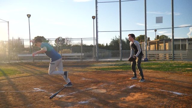 A young man baseball player makes a hit with his bat and runs to first base during a team practice with friends in the park at sunset.